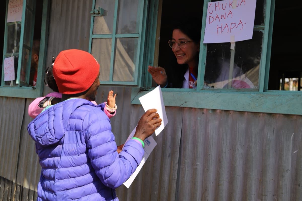 A woman in a purple coat carrying a baby collects paperwork at a camp pharmacy window while a smiling TotalCare volunteer leans out to hand her medication, under a hand-lettered sign reading “Chukua dawa hapa.”