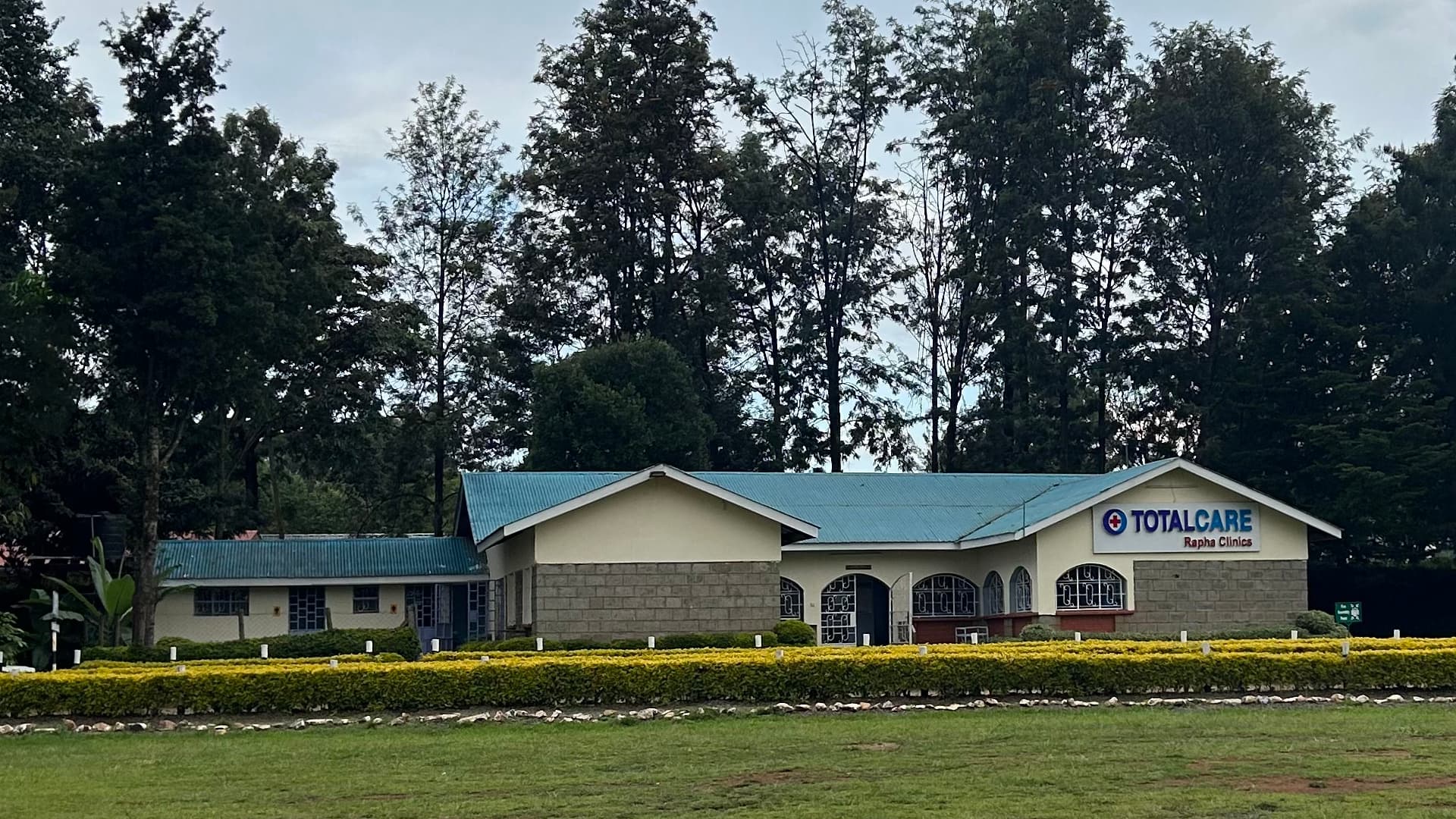 The TotalCare Rapha Clinics building in Eldoret seen across its lawn — a long single-storey block under a blue metal roof, its sign on the near gable, behind a low clipped hedge with tall trees rising behind it.