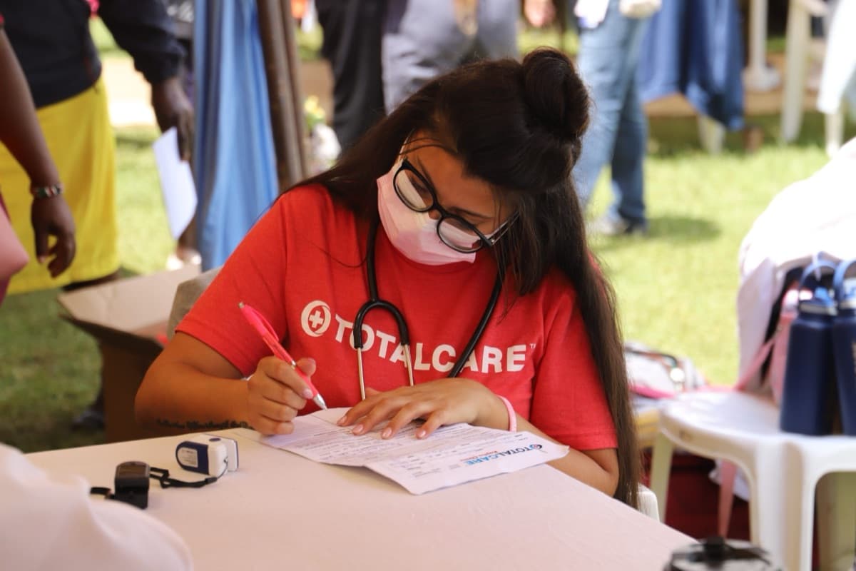 A masked TotalCare volunteer with a stethoscope fills in a patient form at an outdoor camp table.