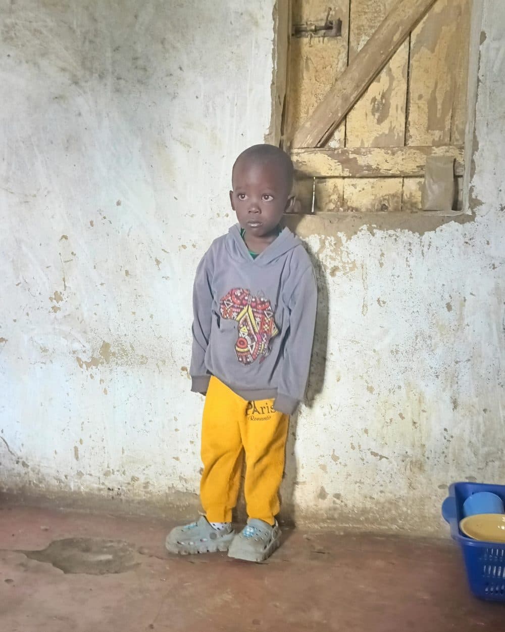 Adrian stands against a plastered wall beneath a wooden window shutter, wearing a grey hooded sweatshirt printed with a patterned map of Africa, bright yellow tracksuit trousers and oversized grey shoes. A blue plastic basket holding a yellow bowl stands on the earth floor at the edge of the frame.