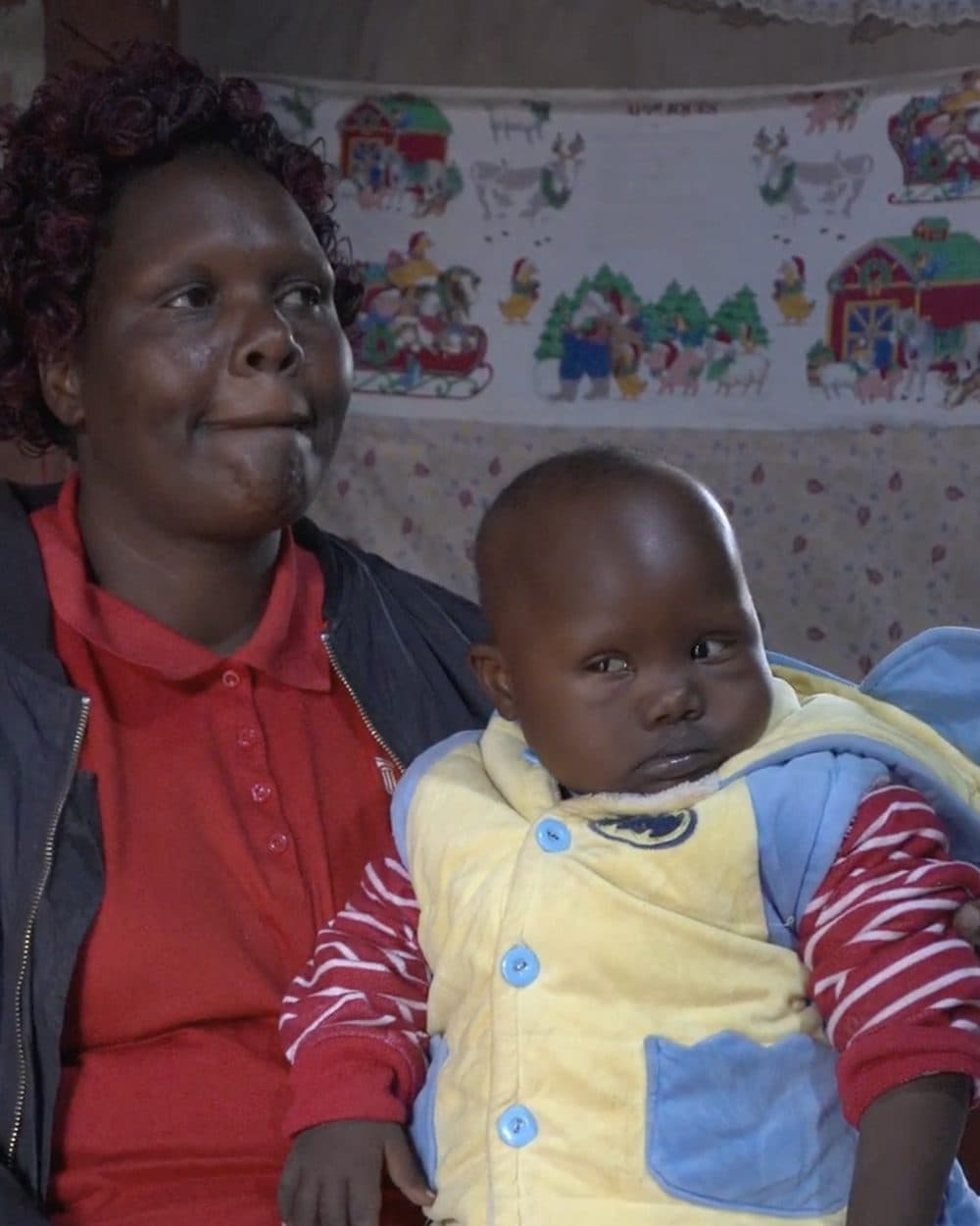 Gianna is held by a woman in a red polo shirt and dark jacket, wearing a padded yellow vest with blue trim over a red and white striped top, her gaze turned to one side. Behind them, a cloth printed with farm scenes hangs against a wall covered in a small floral print.