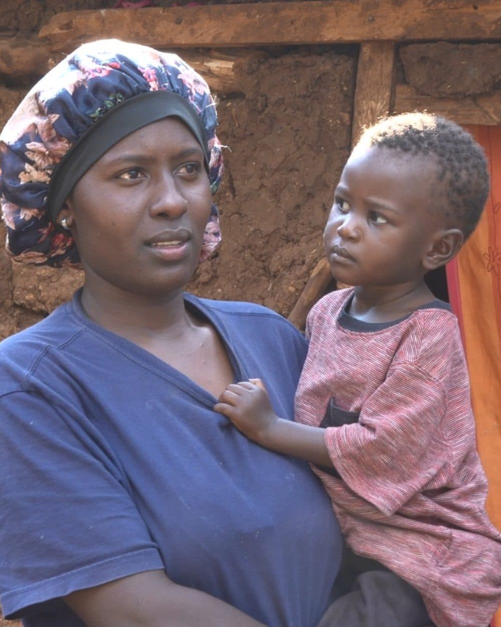 Kelly Ann is carried by a woman in a blue T-shirt and a dark floral bonnet, one small hand holding the front of the woman’s shirt. She wears a red and white striped shirt and looks off to one side; behind them stand an earth-and-timber wall and an orange curtain.