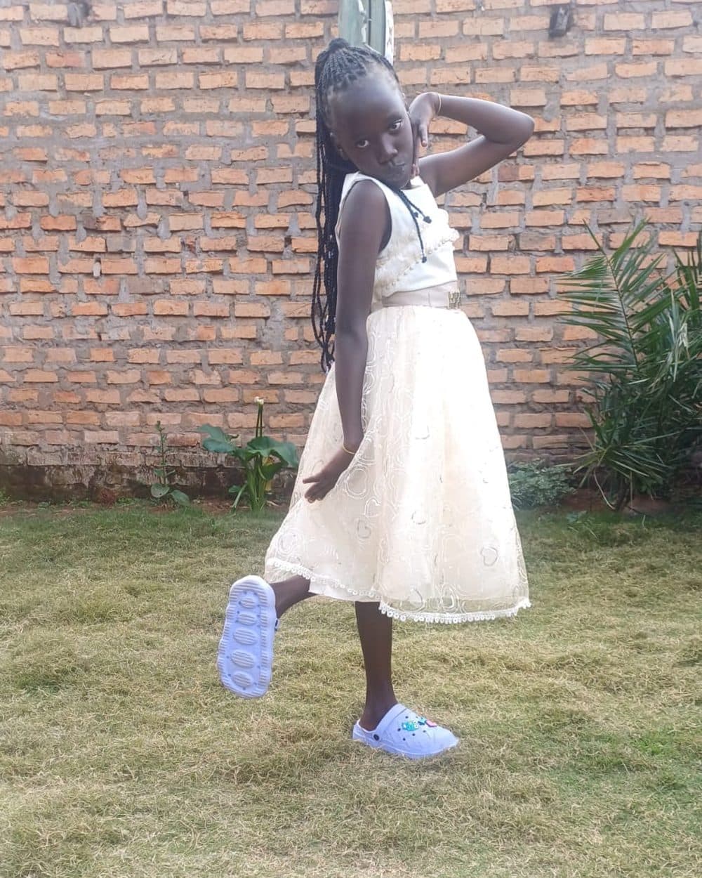 Lorna poses on a lawn in a cream sleeveless dress with a lace hem, long braids falling past her shoulders, one hand raised behind her head and one foot kicked up behind her, light blue clogs on her feet. A brick wall and a green shrub stand behind her.