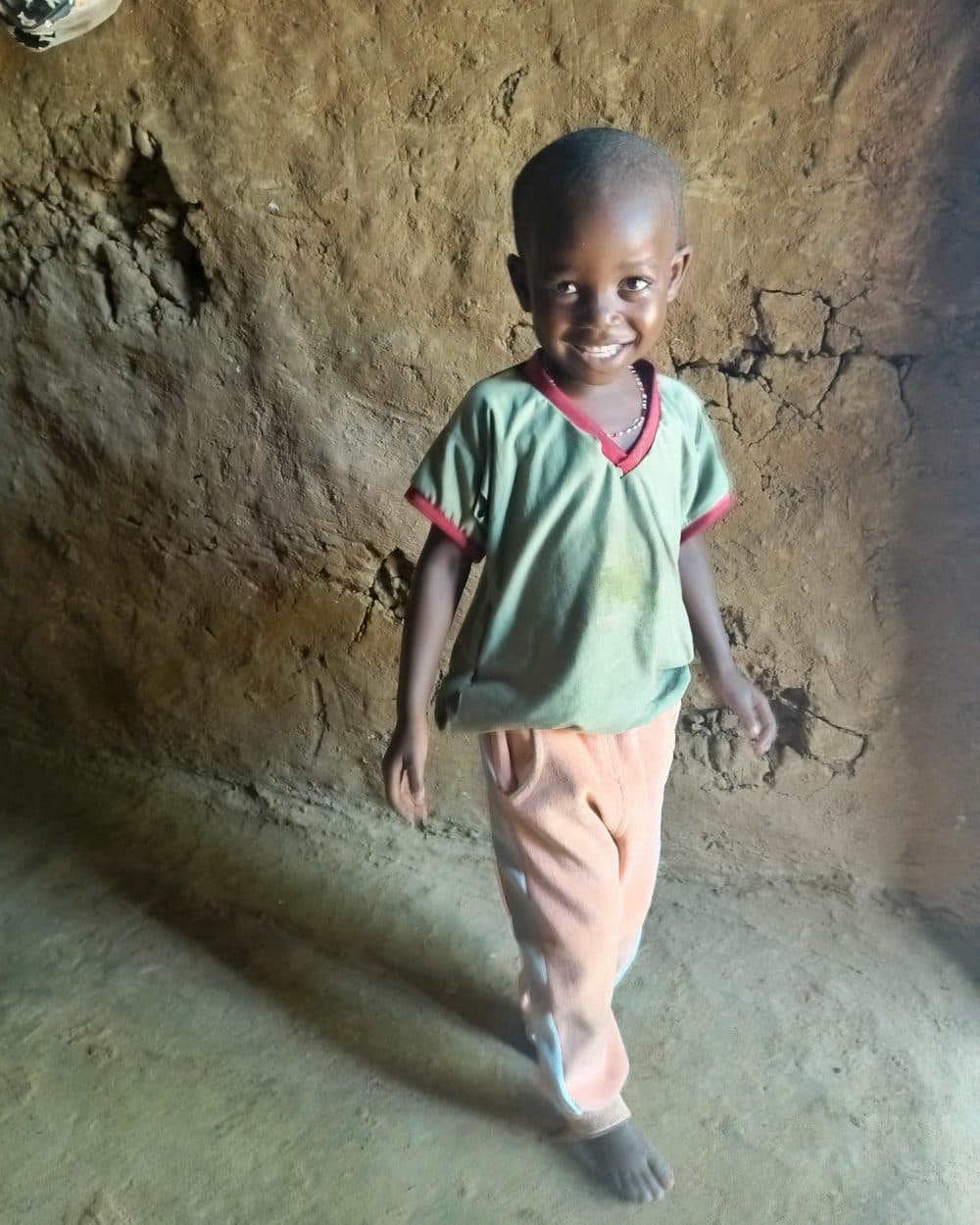 Princess walks towards the camera, smiling, in a green short-sleeved top with red trim at the collar and cuffs and a thin beaded necklace, pink trousers, and bare feet on a swept earth floor. An earth wall stands behind her.
