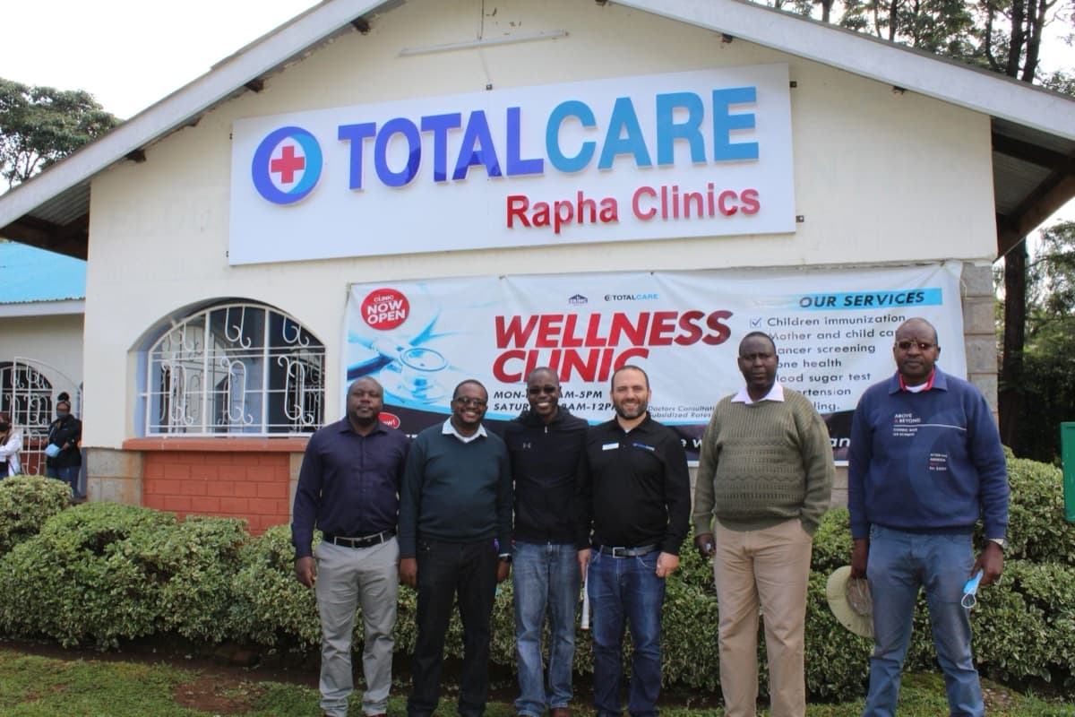 Six men stand outside the clinic beneath the TotalCare Rapha Clinics sign and a Wellness Clinic banner listing the services offered.