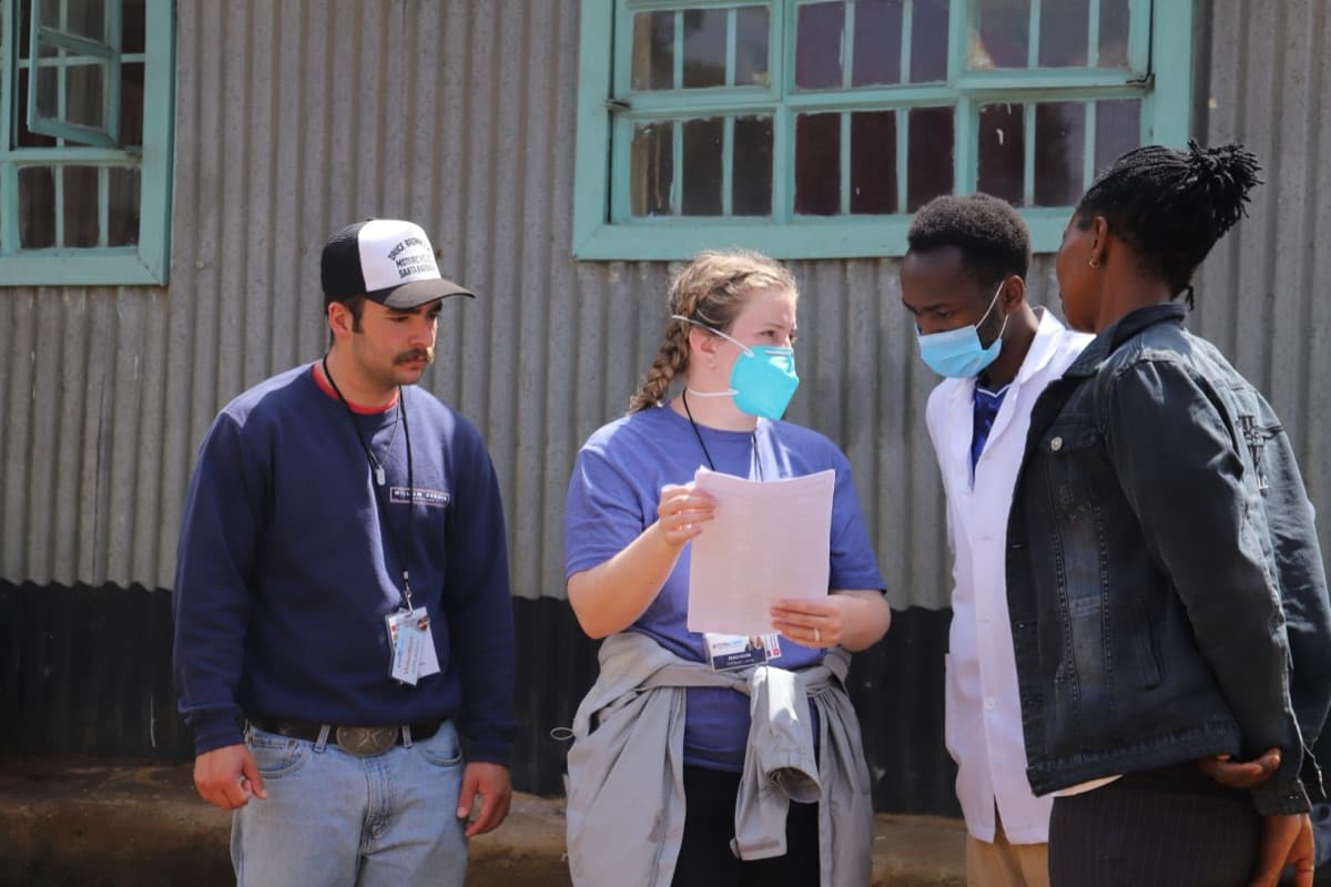 Four people talking outside a corrugated-metal building: two badged TotalCare volunteers, one holding a patient form, a masked clinician in a white coat, and a woman in a denim jacket.