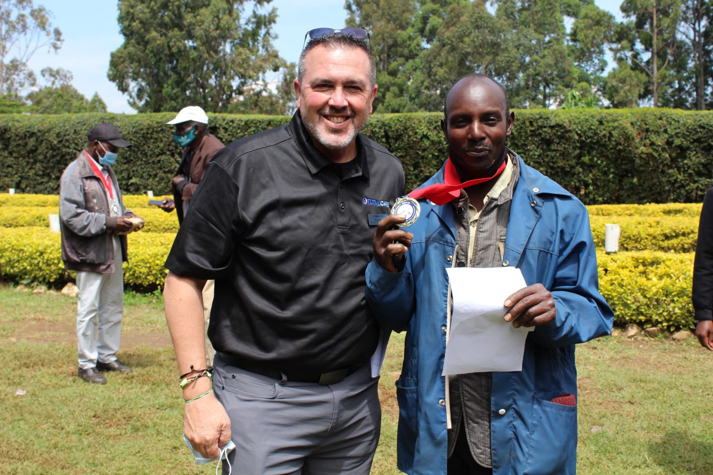 A TotalCare staff member in a black polo stands arm in arm on the grass with a man in a blue jacket and red scarf who holds up a gold medal and a sheet of paper; masked visitors and trimmed hedges behind them.