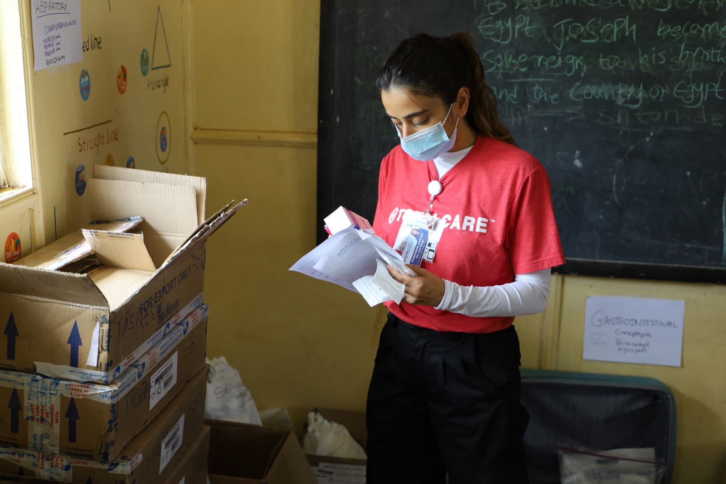A masked TotalCare volunteer in a red shirt reads a medication chart beside stacked supply boxes in a classroom set up as a camp pharmacy.
